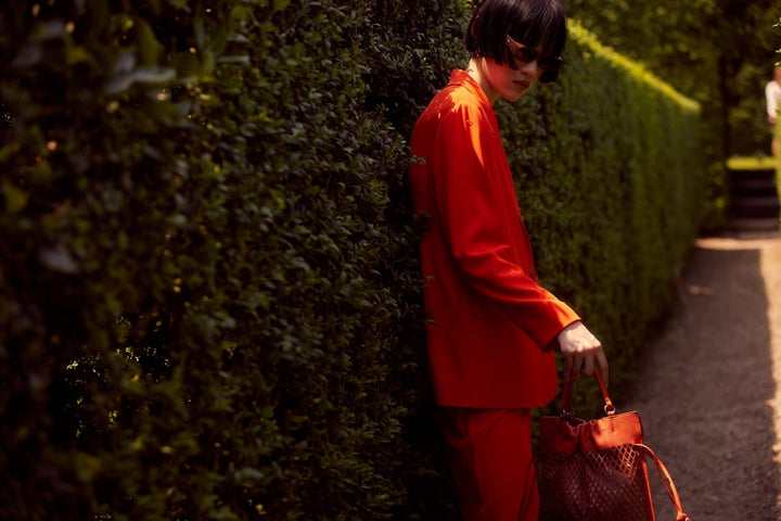 Person in a fashionable red suit and sunglasses standing by a leafy green hedge