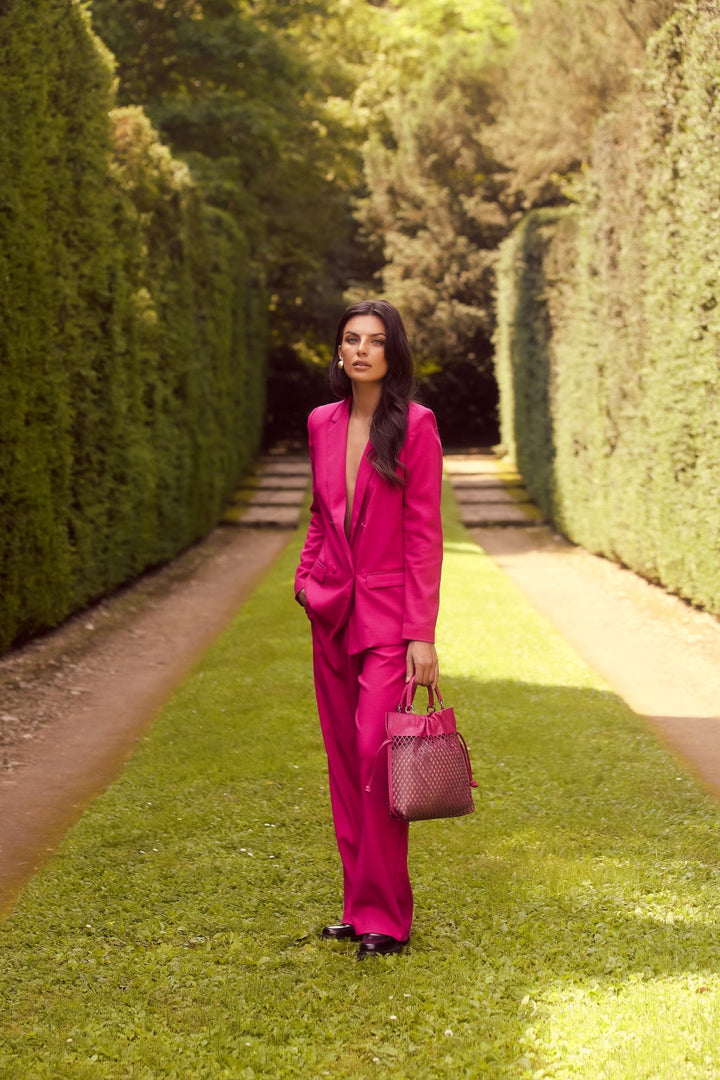 Woman in a bright pink suit standing on a garden path lined with tall hedges