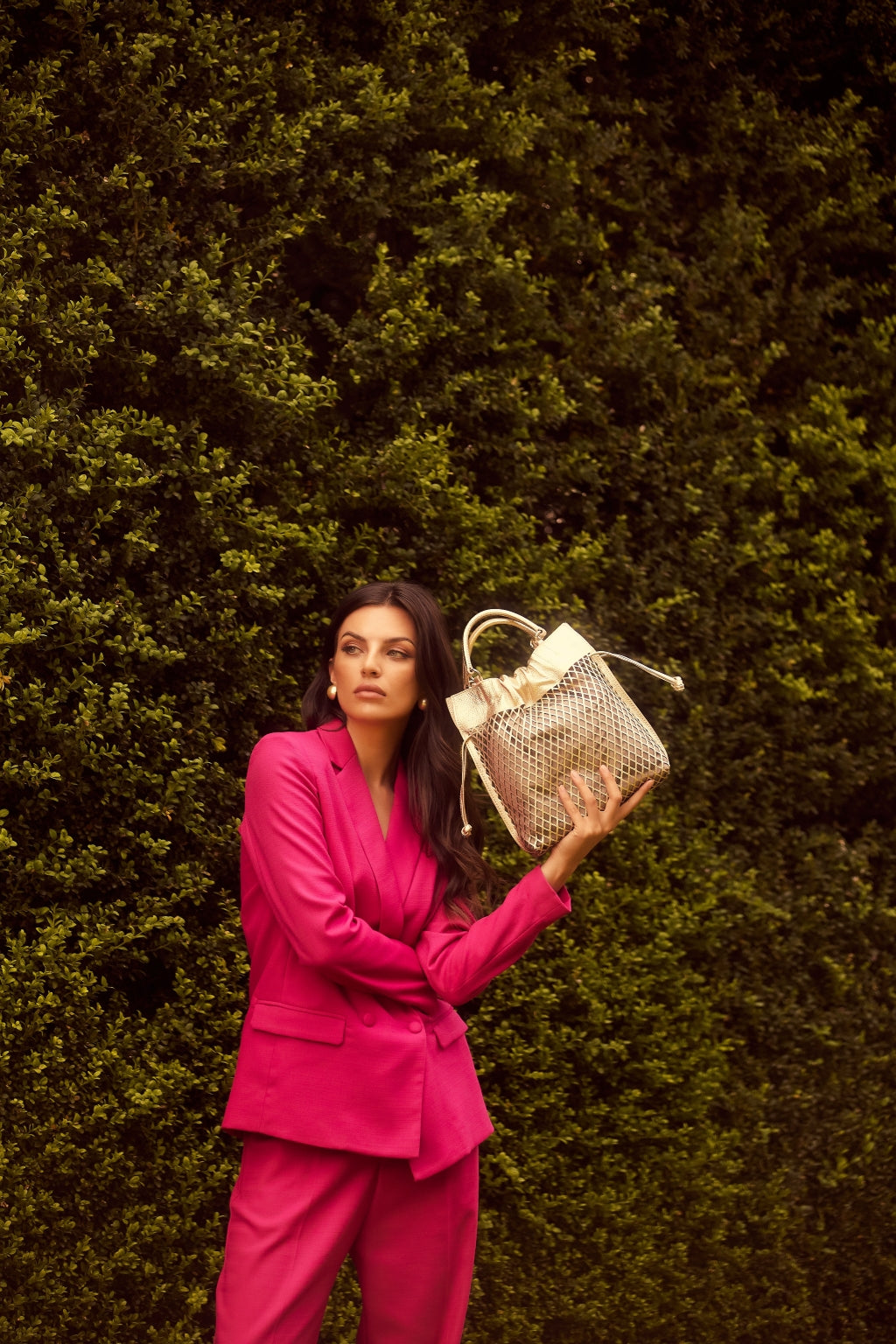 Woman in vibrant pink suit holding a designer handbag against a lush green backdrop