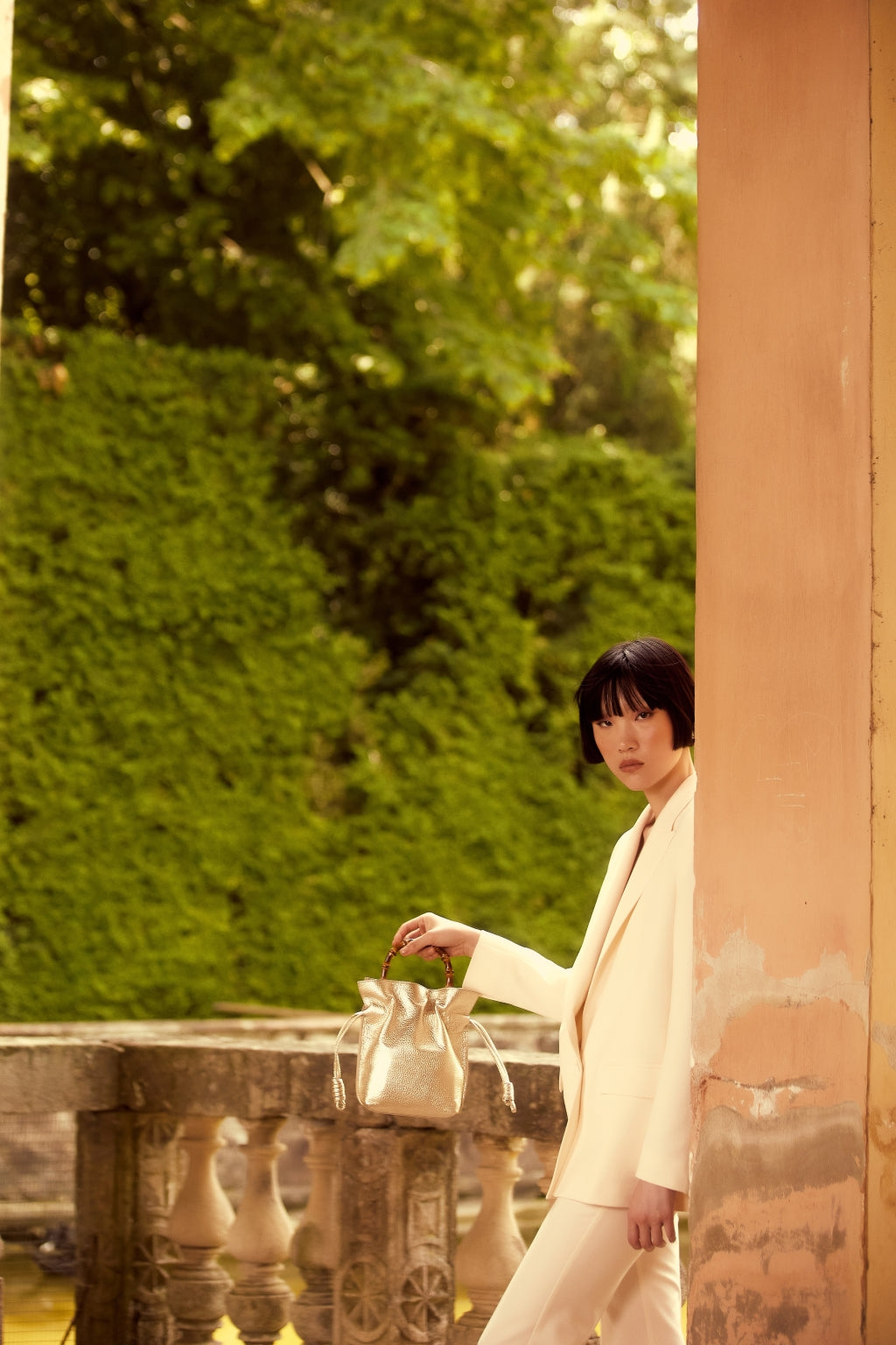 Woman in white suit holding a gold handbag in a leafy garden setting