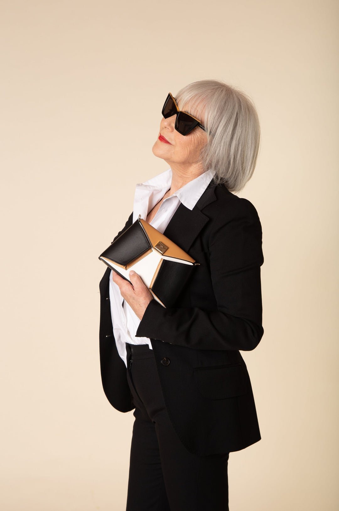 Stylish elderly woman in black suit and sunglasses holding a clutch against a beige background