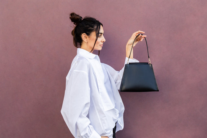 Woman with a bun hairstyle in a white blouse holding a black handbag against a pink background