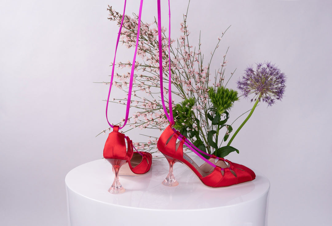 Red high-heeled shoes with transparent heels displayed on white pedestal, surrounded by pink and green flowers