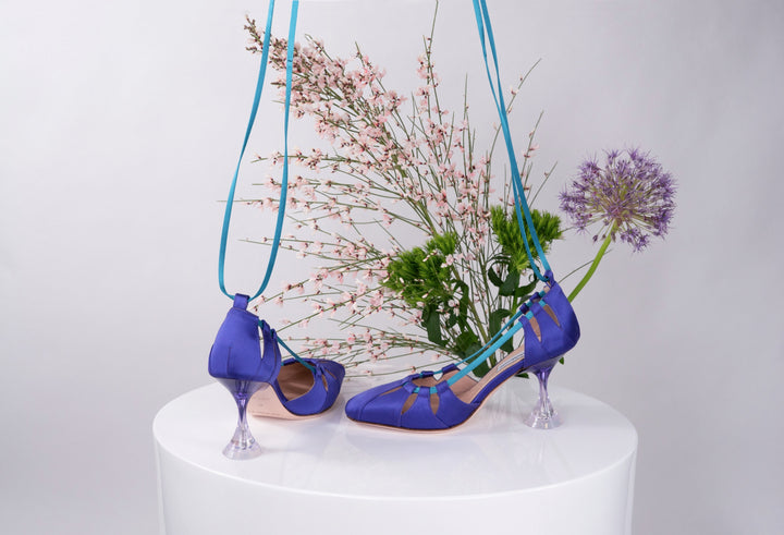 Purple high-heeled shoes with transparent heels against a floral backdrop on a white surface