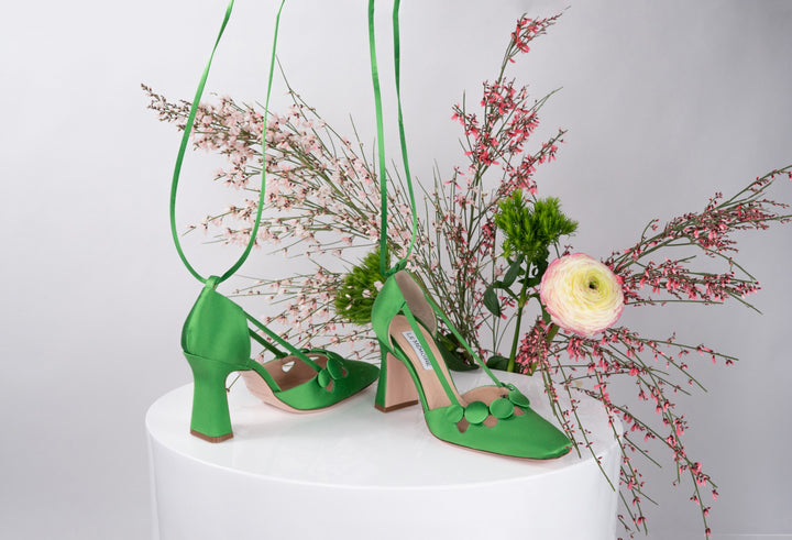 Green high-heeled shoes with flower decorations on a white pedestal surrounded by pink flowers
