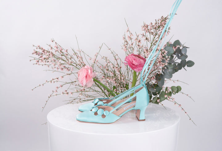 Blue high-heeled shoes with ankle straps on a white pedestal, adorned with pink and green flower arrangement