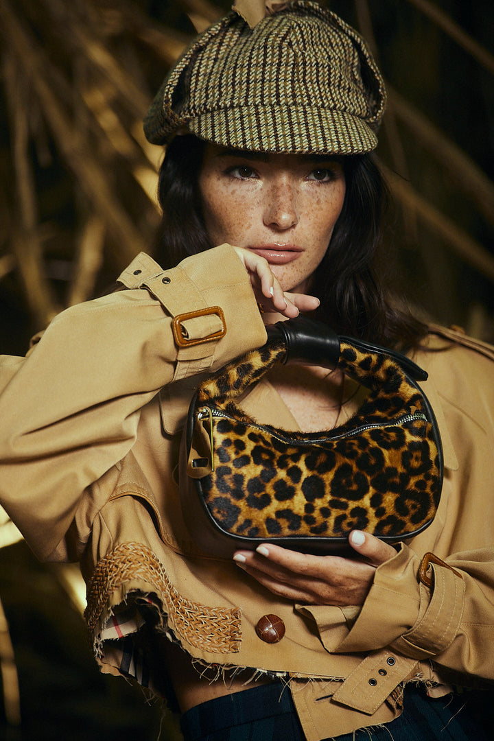 Woman wearing a plaid hat, beige jacket, and holding a leopard print handbag in an artistic outdoor setting