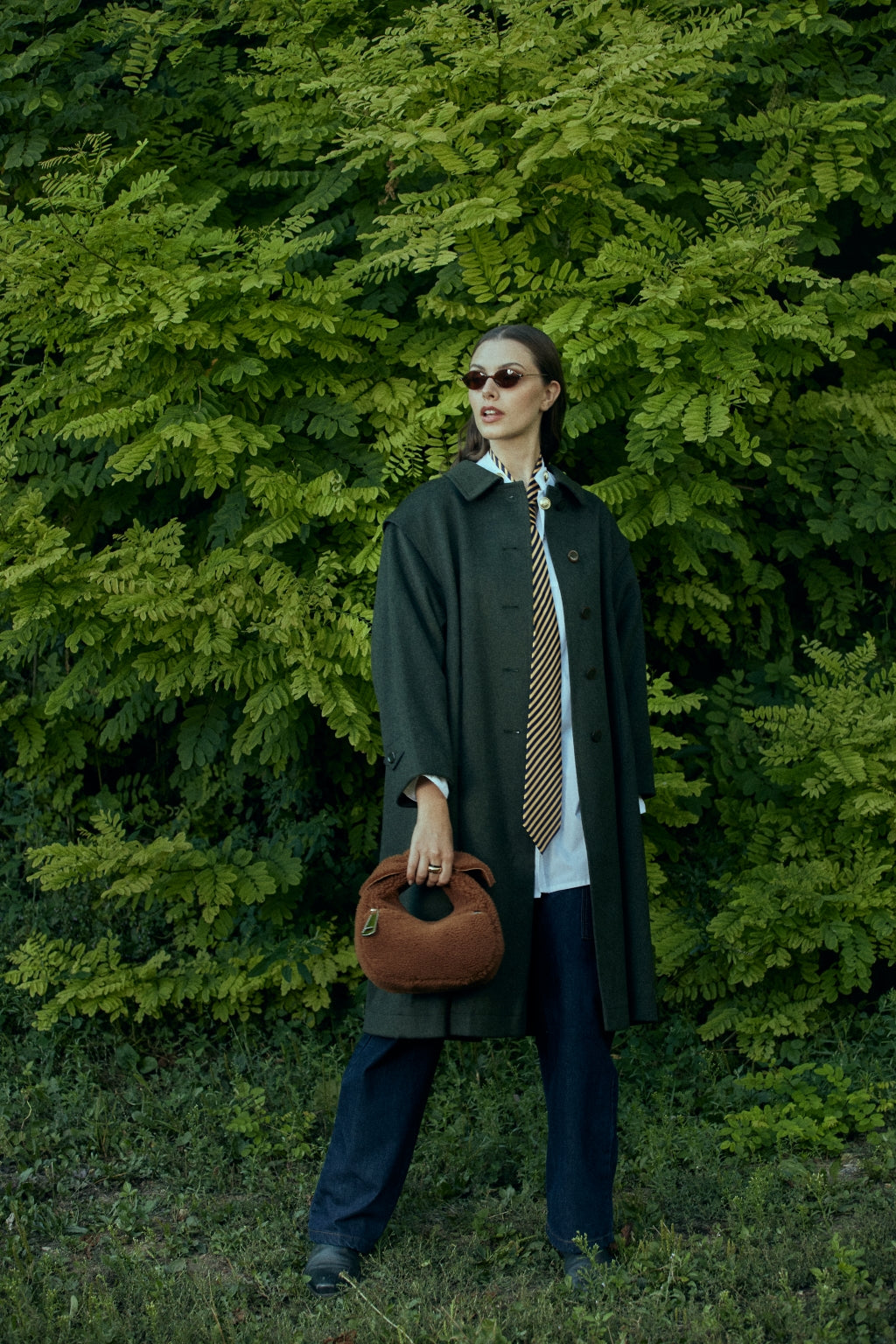 Woman in stylish oversized coat, sunglasses, and tie holding a handbag in front of lush green foliage