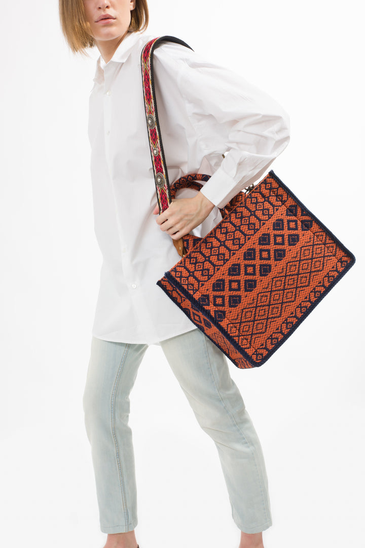 Person wearing a white shirt and light jeans, carrying a large red and blue patterned bag with an ornate strap