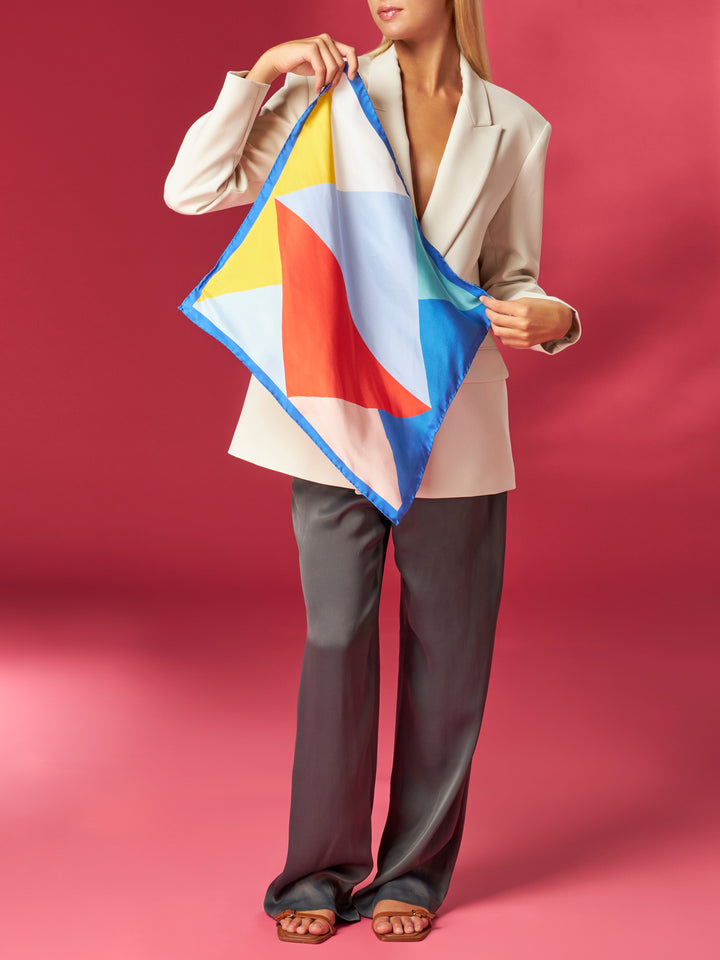 Woman in a stylish white blazer and gray trousers showcasing a colorful geometric patterned scarf on a red background