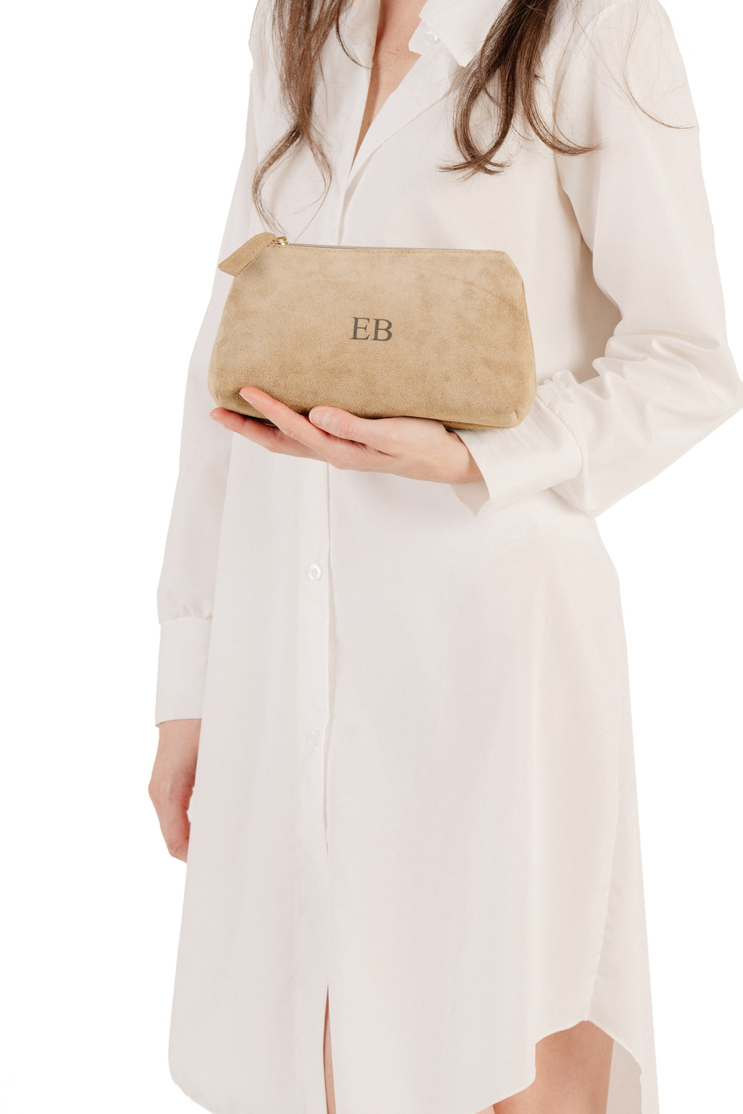 Woman holding tan suede cosmetic bag with initials EB embroidered, wearing white shirt dress