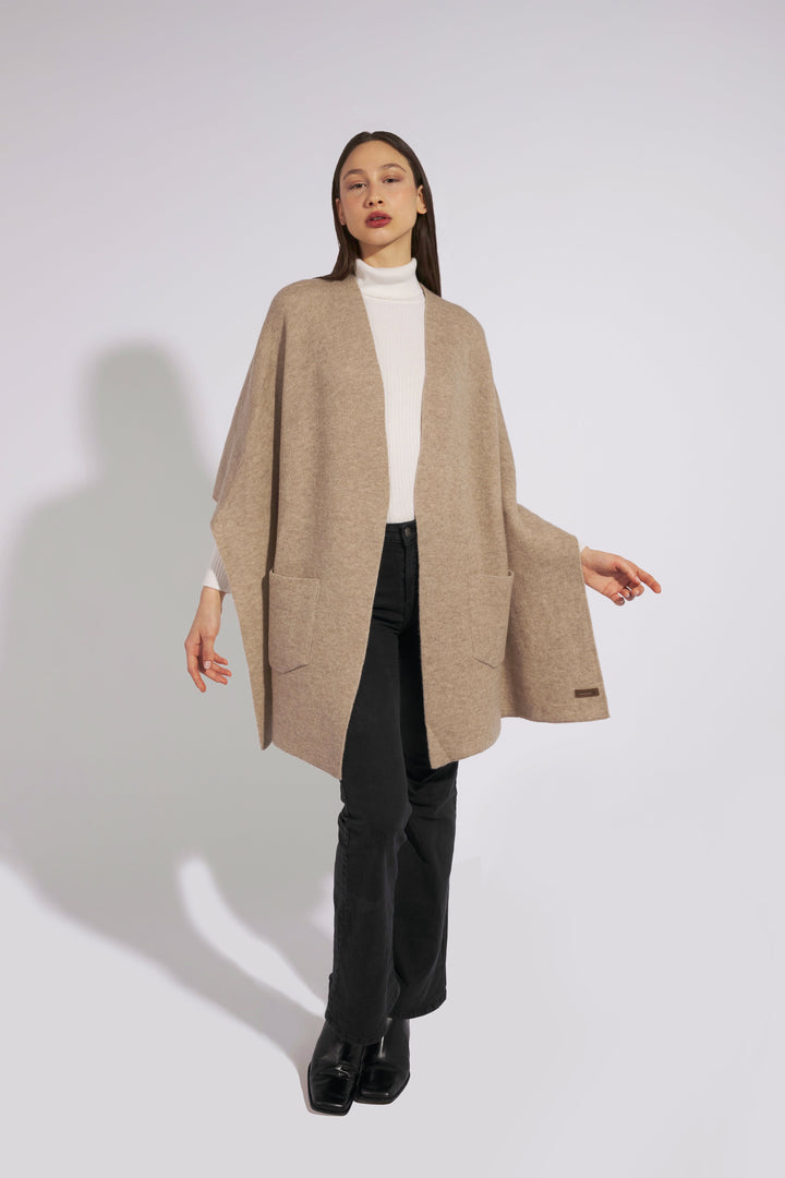 Woman wearing beige cardigan over white turtleneck and black pants against plain background