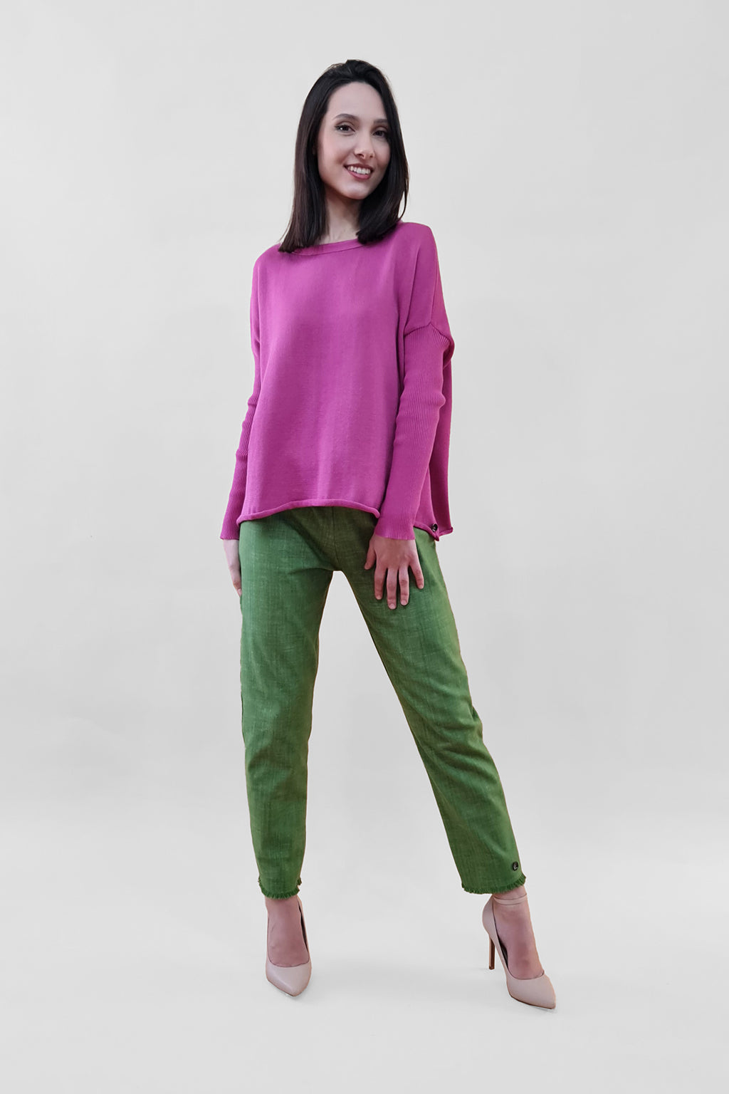 Woman wearing a pink sweater with green pants and beige heels standing against a plain background