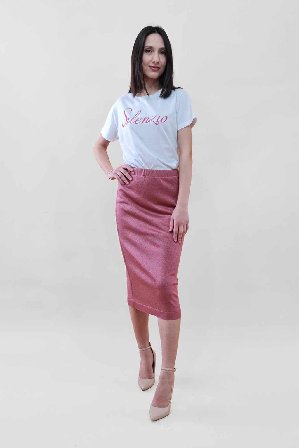 Woman in a white Silenzio t-shirt and pink skirt with beige heels standing against a white background