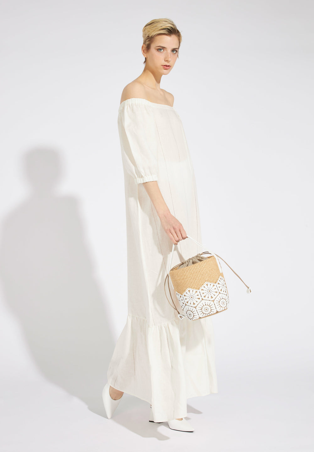 Woman wearing an off-the-shoulder white dress with a wicker handbag and white heels against a white background