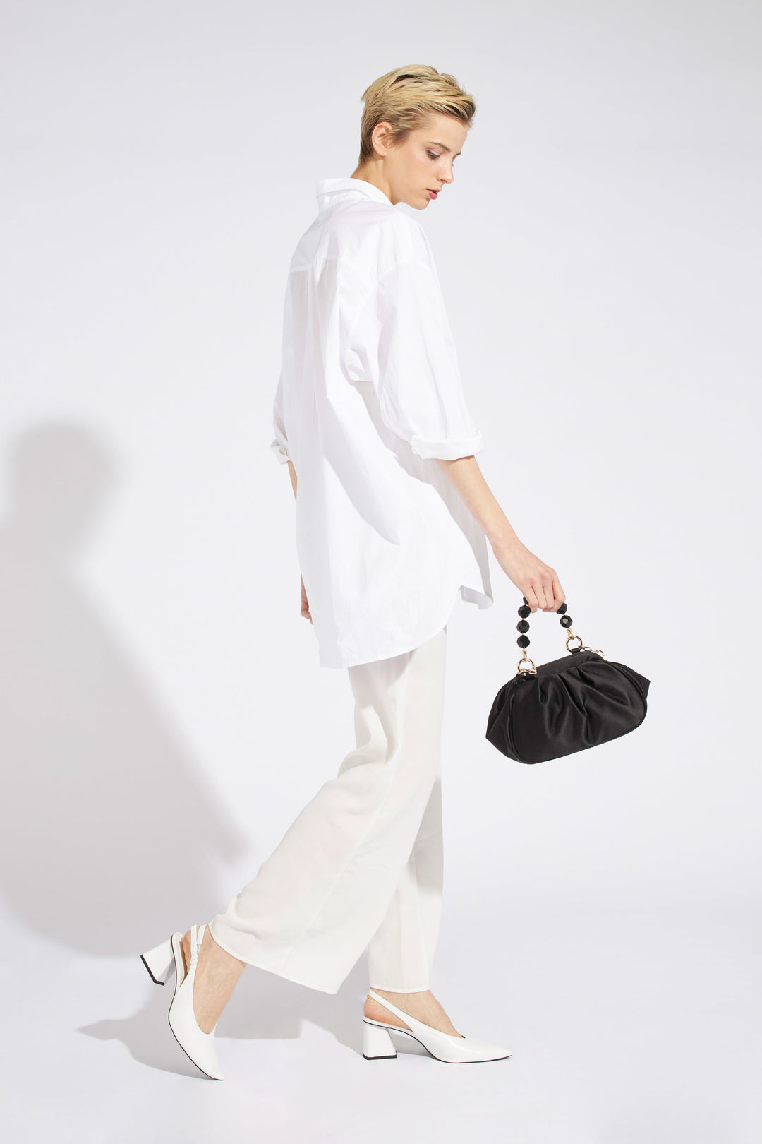 Fashion model in white oversized shirt and pants holding a black handbag and wearing white heels