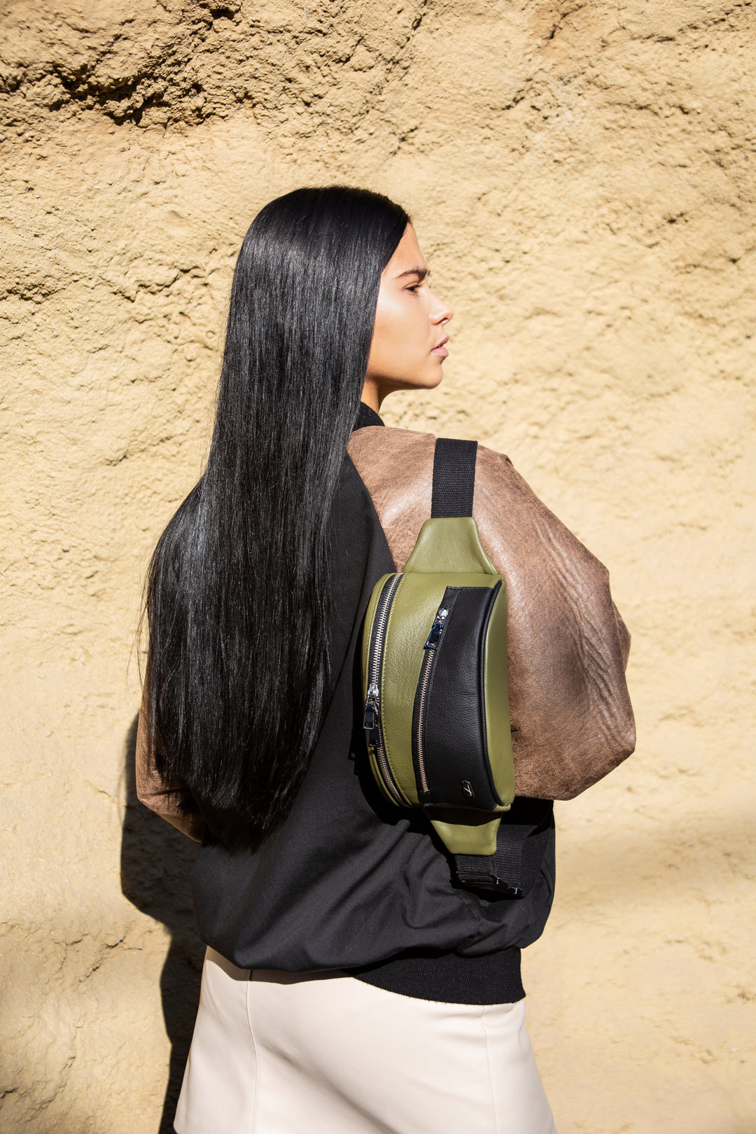 Woman with long black hair wearing a green and black backpack standing against a beige textured wall