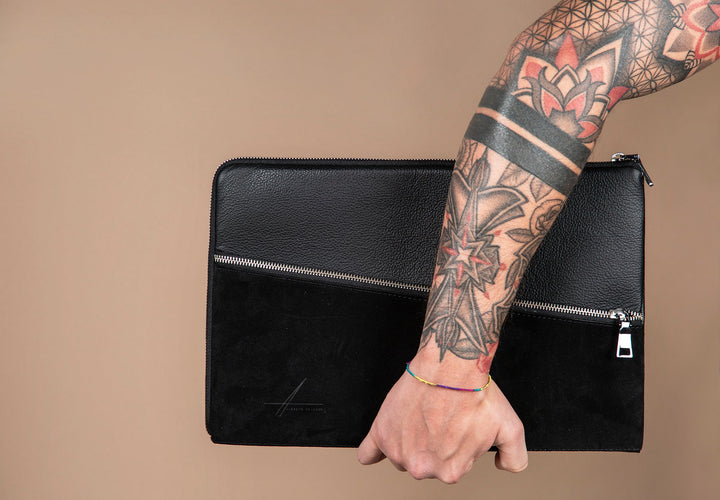 Person holding black leather laptop sleeve with visible tattoos on arm against beige background