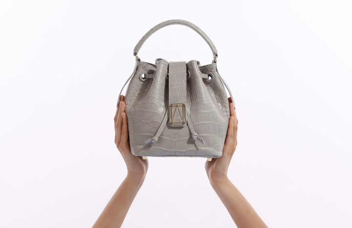 Hands holding a gray crocodile-patterned leather handbag against a white background