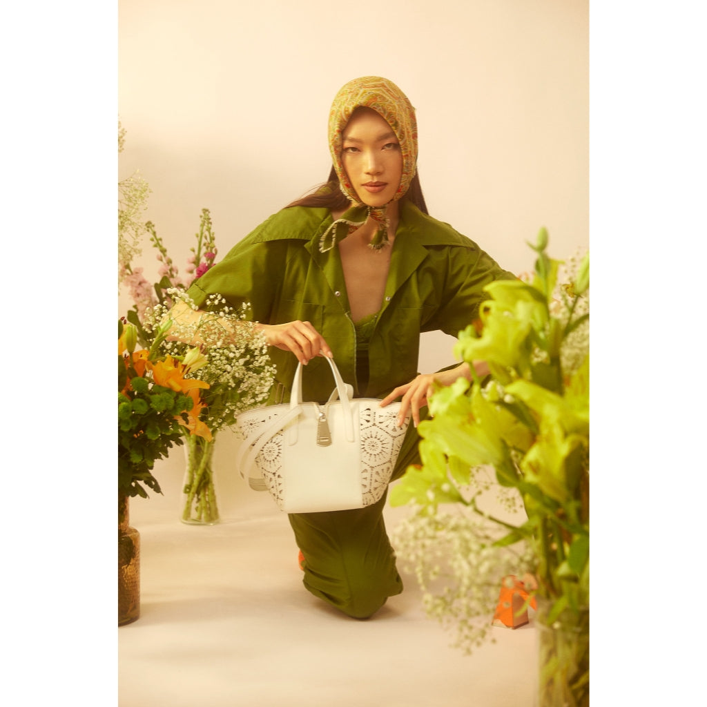 Woman in green outfit with a patterned headscarf, holding a white lace handbag, surrounded by colorful flowers