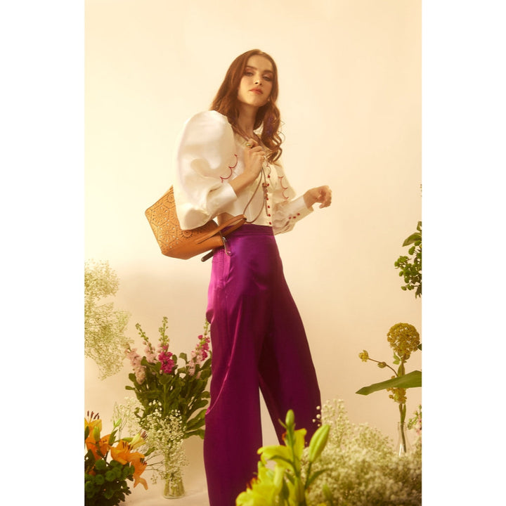 Fashionable woman in vibrant purple pants and white blouse with puffy sleeves, surrounded by flowers and plants
