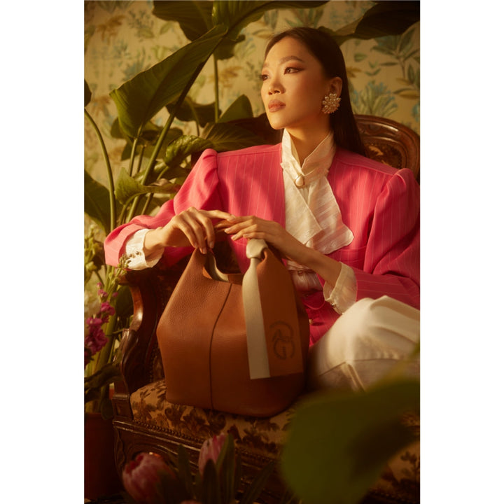 Woman in elegant pink blazer sitting indoors with a brown handbag in a lush, plant-filled setting