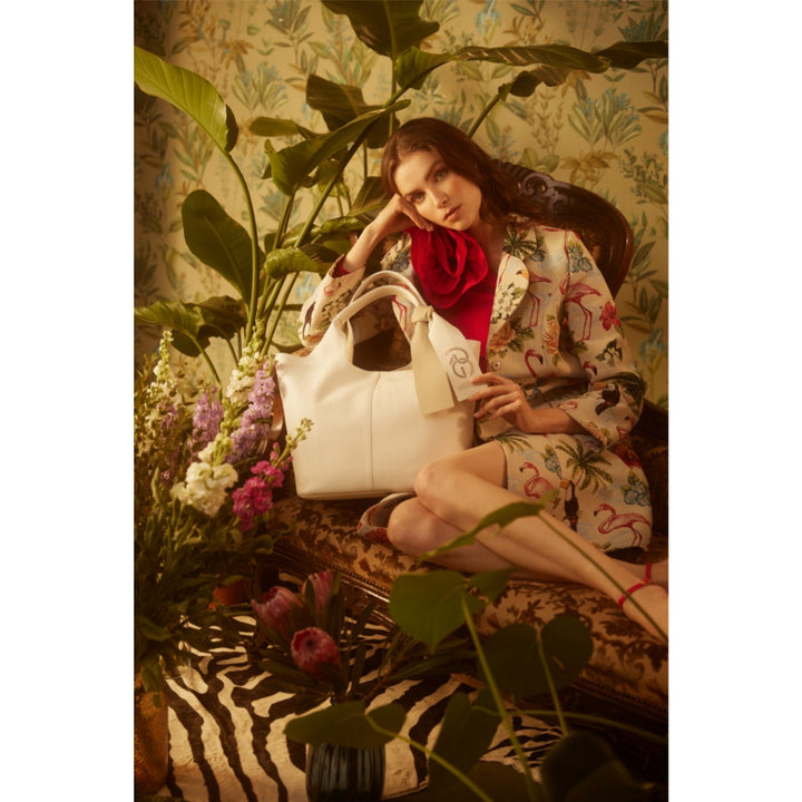 Woman sitting in decorated room with floral wallpaper, holding a white handbag, surrounded by plants and flowers