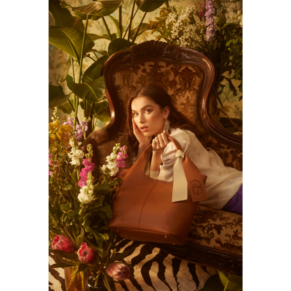 Woman in vintage chair surrounded by flowers, holding brown leather handbag