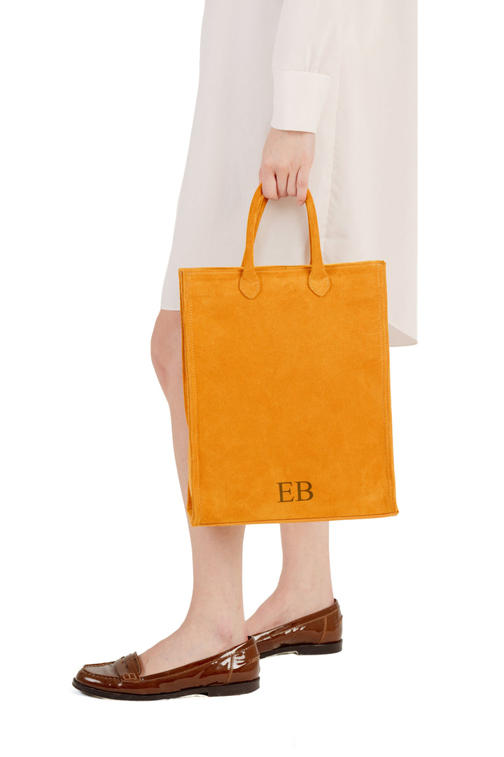 Person holding a vibrant orange suede tote bag with initials EB wearing a white dress and brown patent leather loafers