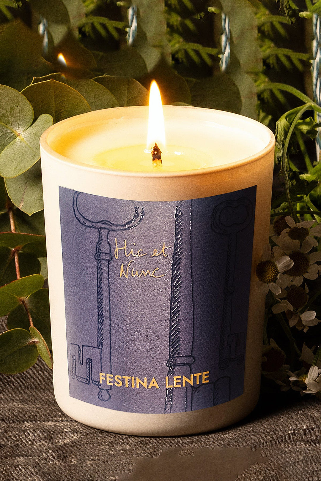 Lit candle in a white holder with Festina Lente label, surrounded by greenery