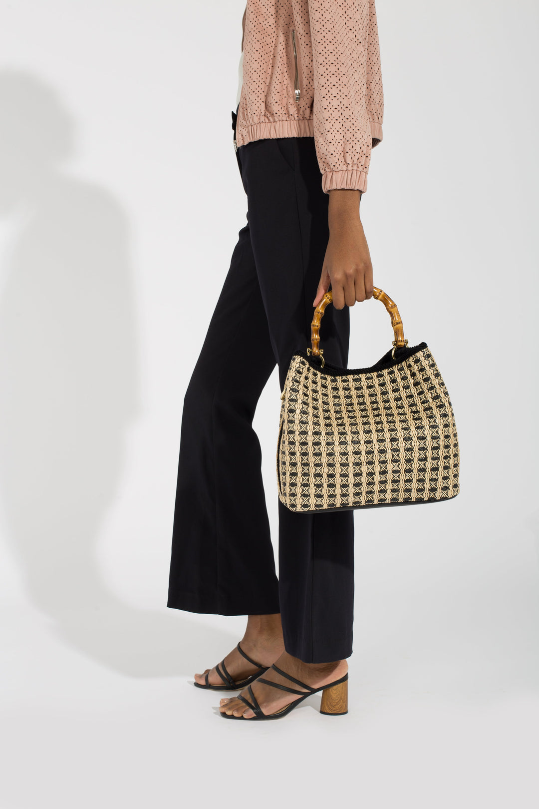 Woman holding a woven handbag with bamboo handles while wearing black pants and heeled sandals