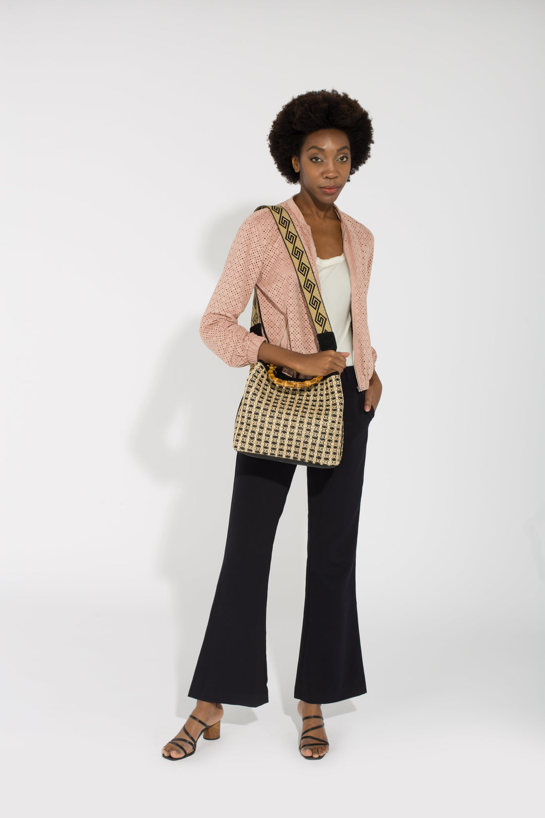 Woman in stylish attire with pink blazer, white top, black flared pants, and patterned shoulder bag, standing against white backdrop