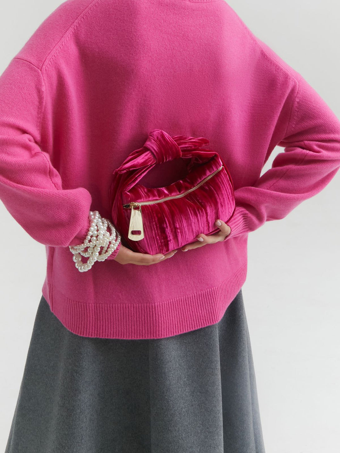 Person holding pink velvet clutch bag and wearing a bright pink sweater and pearl bracelet