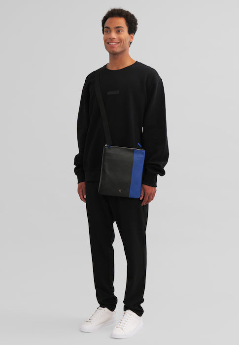 Man in black casual outfit with black and blue crossbody bag on gray background