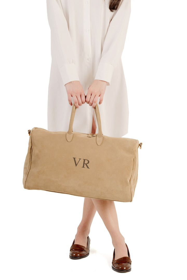 Woman holding tan suede duffle bag with initials 'VR' in front of white background