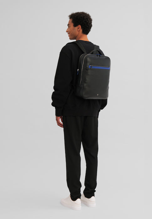 Person wearing a black outfit and a backpack, standing against a gray background
