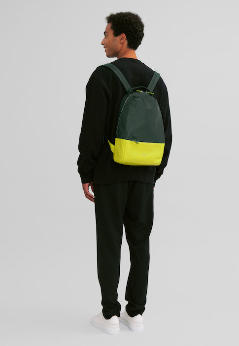 Man wearing black outfit and green-yellow backpack against light gray background