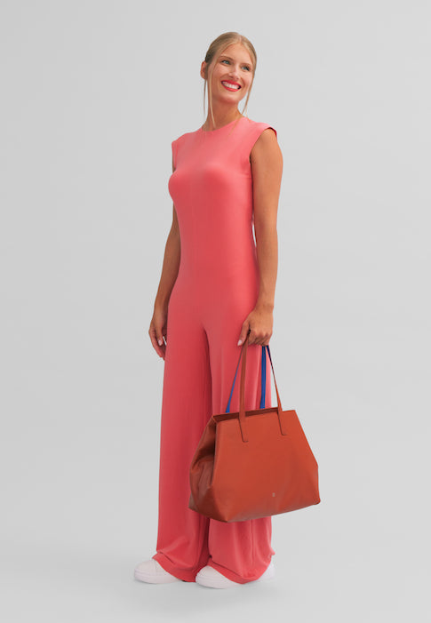 Smiling woman in a pink jumpsuit holding an orange handbag against a gray background