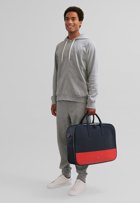 Man wearing a grey tracksuit holding a navy and red bag in front of a plain background