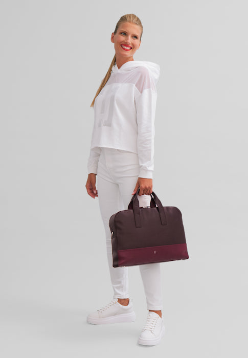 Woman wearing white outfit and holding a maroon and dark brown handbag against a plain background