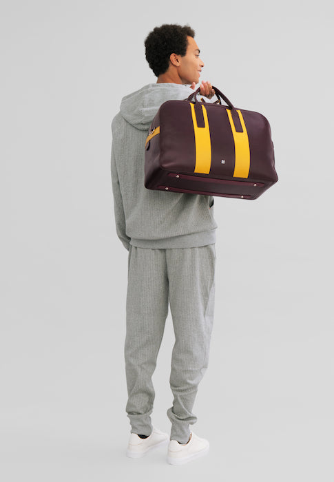 Person in gray tracksuit carrying a large maroon and yellow duffel bag over shoulder