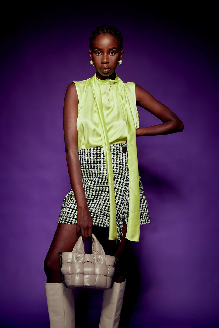 Fashionable woman in lime green blouse, black-and-white checkered skirt, holding a beige handbag, standing against a purple background