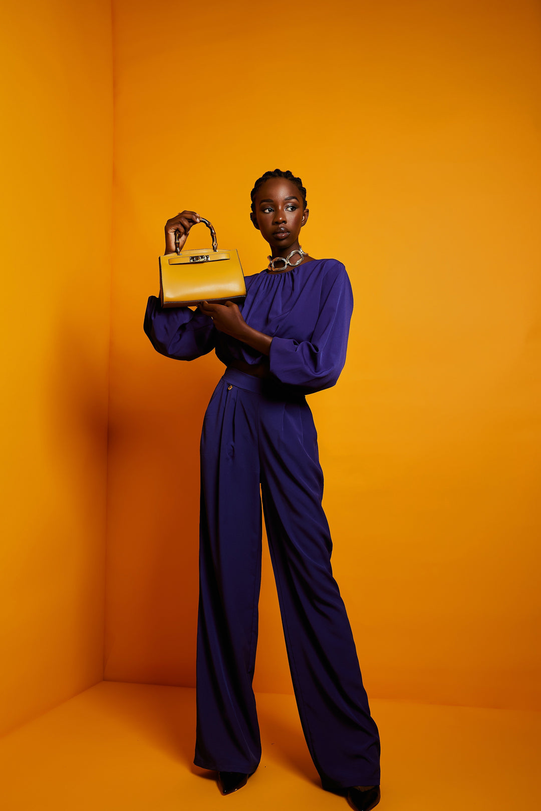 Model in purple outfit holding a yellow handbag against orange background