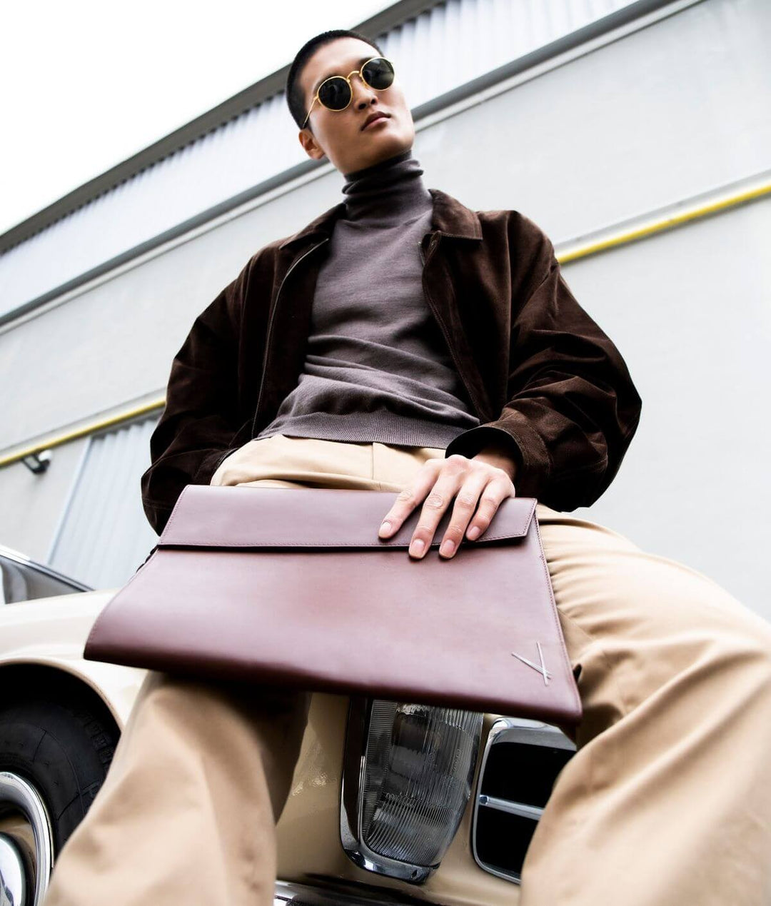 Fashionable person holding a leather portfolio case, wearing sunglasses, standing in front of a vintage car