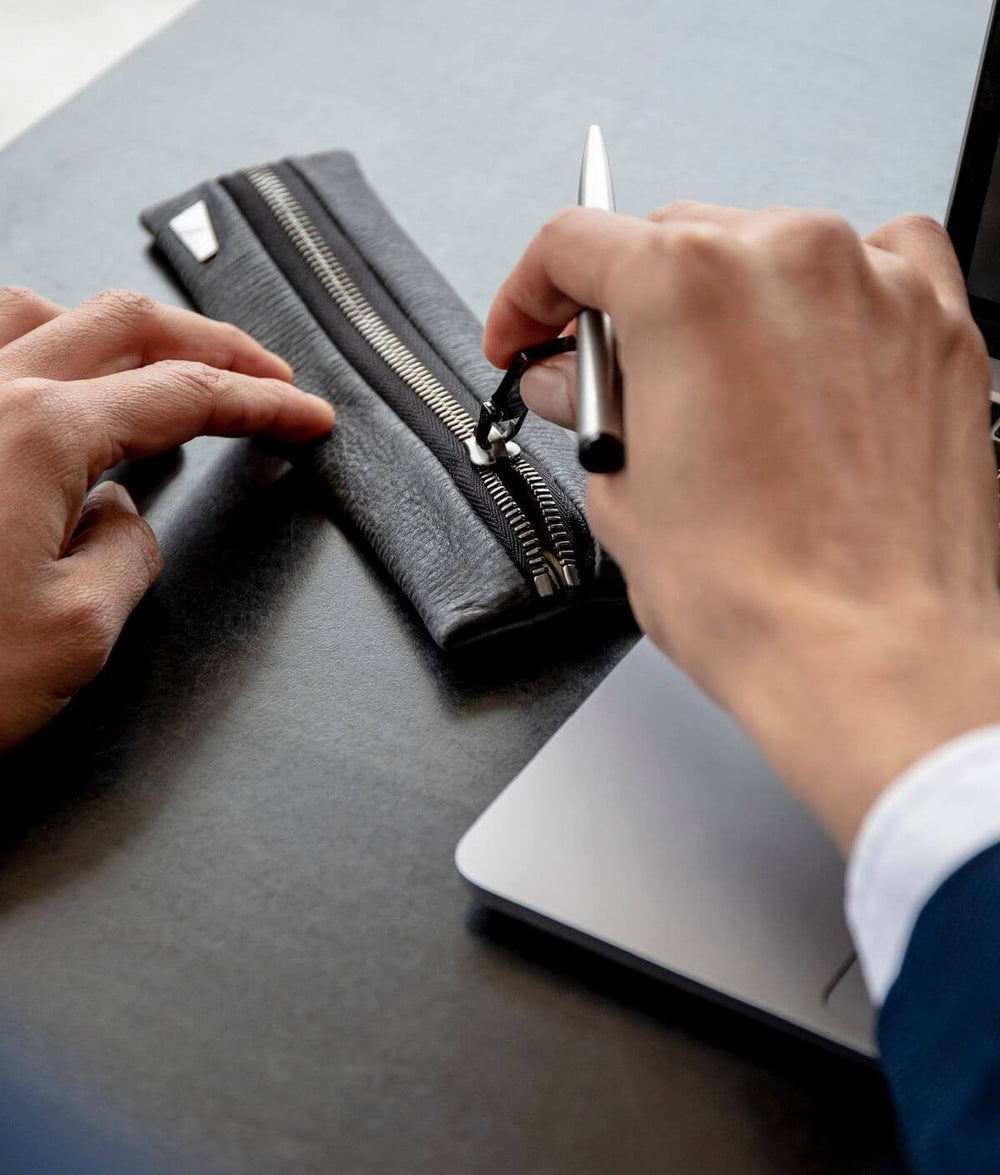 Person opening a black leather pencil case with a zipper beside a laptop