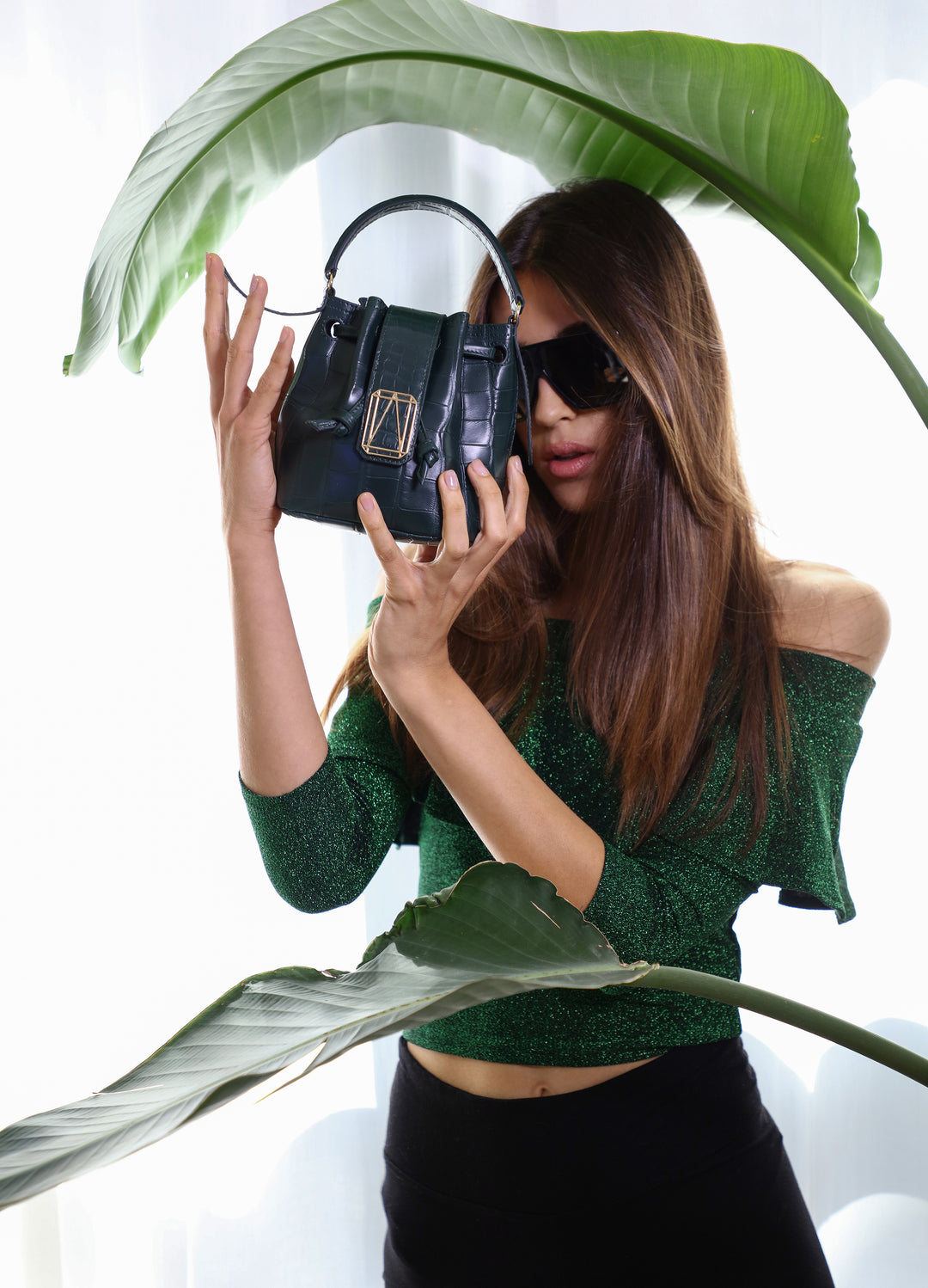 Woman in green outfit and sunglasses holding stylish handbag among large leaves
