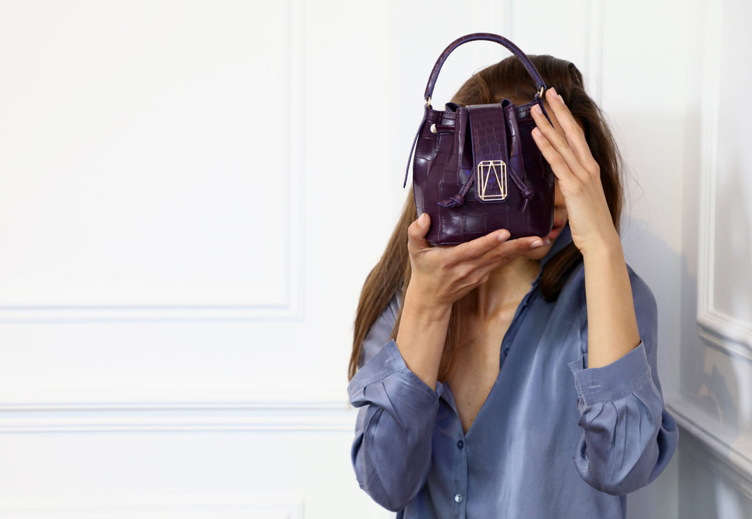 Woman holding a small purple handbag in front of face against white background