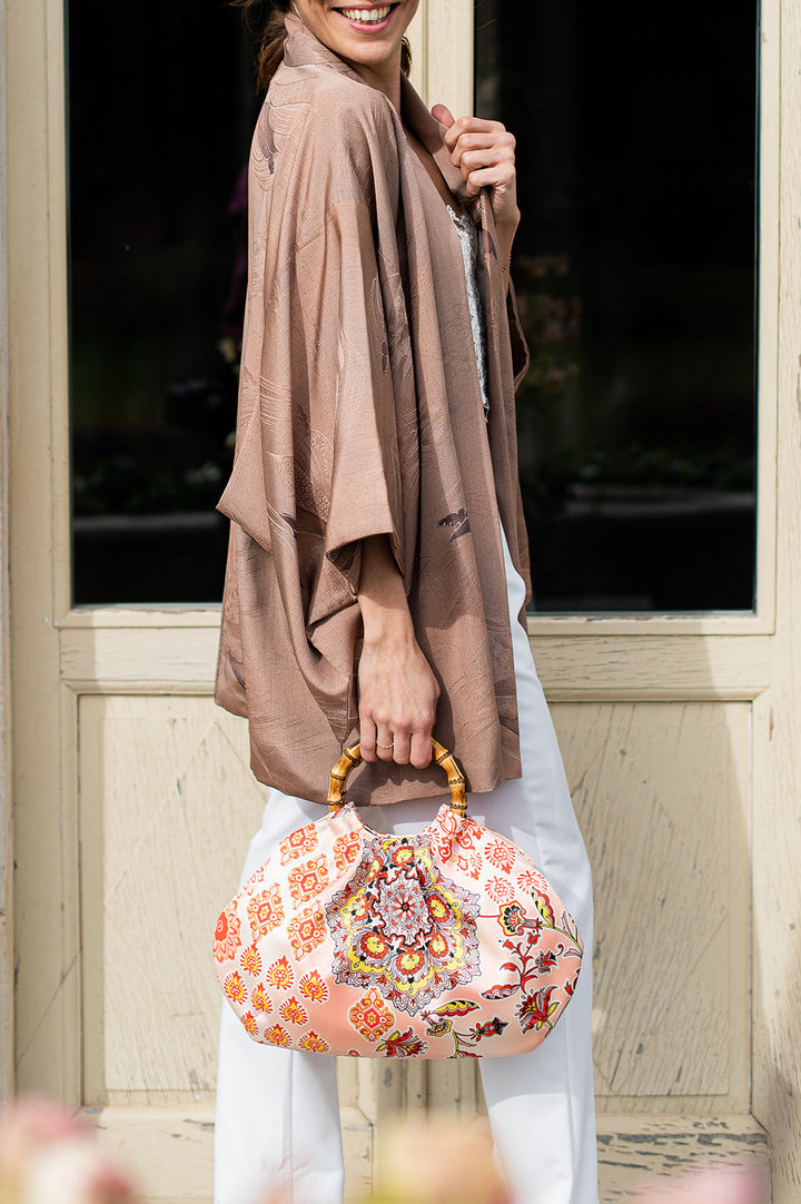 Woman wearing a brown kimono-style jacket and white pants holds a floral-patterned handbag with bamboo handles