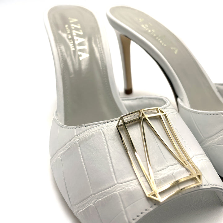 White high-heeled mules with embossed texture and a gold geometric buckle on the front