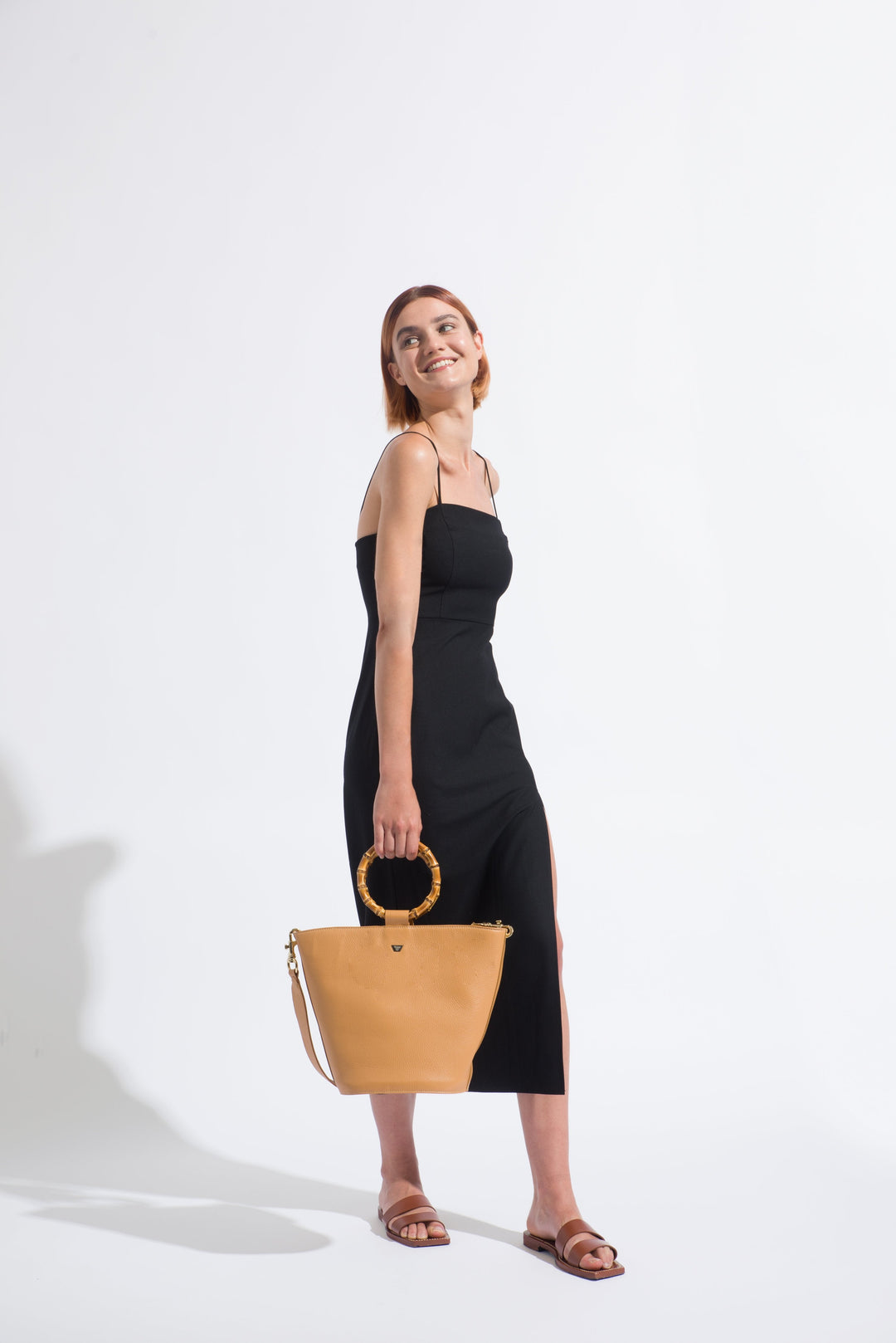Woman in black sleeveless dress holding tan handbag against white background